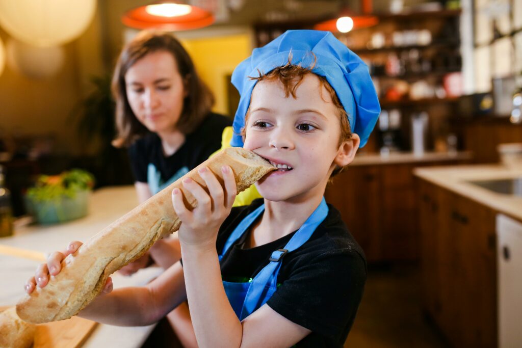 A cheerful child wearing a chef's hat enjoys a homemade baguette in a cozy kitchen setting.